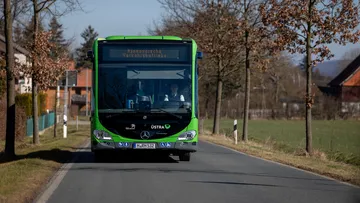 Ein ÜSTRA Bus fährt über eine Landstraße. Zu sehen ist der Bus von vorne. Im Hintergrund sind Bäume, Häuser und Felder zu sehen.