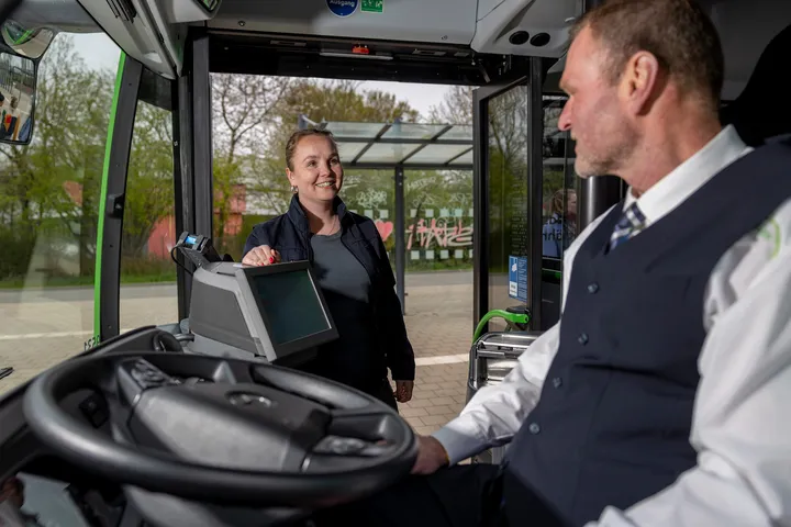 Fahrer im Bus sitzend spricht mit einsteigendem weiblichem Fahrgast 