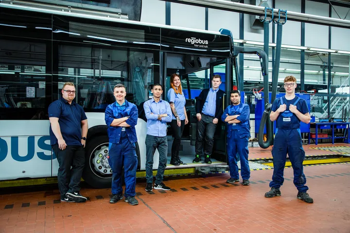Sieben Azubis stehen im Einstiegsbereich eines Busses und davor in der Werkzeughalle
