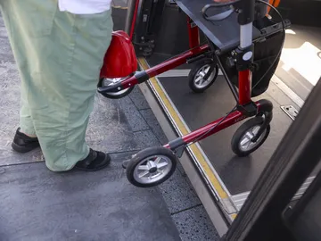 Nahaufnahme auf eine Person, die mit einem Rollator in einen Bus einsteigt. Die Perspektive ist von oben, man sieht die Räder des Rollators und die Beine der Person.  