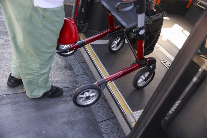 Nahaufnahme auf eine Person, die mit einem Rollator in einen Bus einsteigt. Die Perspektive ist von oben, man sieht die Räder des Rollators und die Beine der Person.  