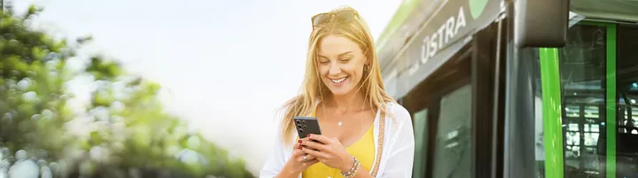 Frau steht vor einem Bus und schaut auf ihr Smartphone, dabei lacht sie