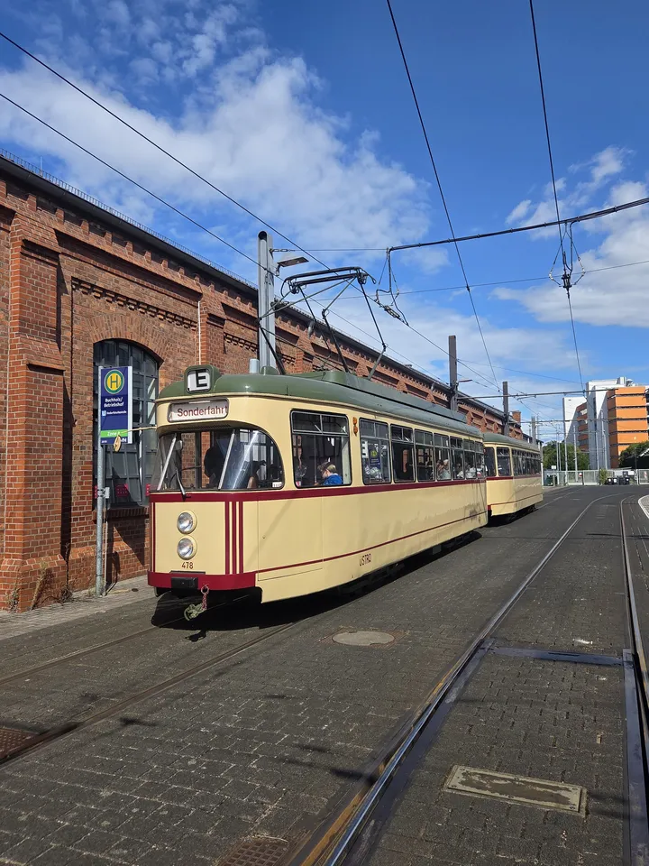Eine Oldtimer-Stadtbahn in beige und dunkelrot steht auf Gleisen vor einem Backsteingebäude.