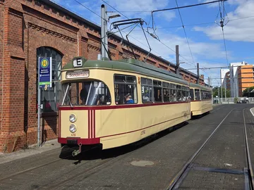 Eine Oldtimer-Stadtbahn in beige und dunkelrot steht auf Gleisen vor einem Backsteingebäude.