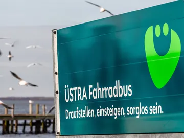 Ein Rennrad links im Bild, der ÜSTRA Fahrradbus-Anhänger rechts im Bild, im Hintergrund sieht man das Steinhuder Meer und einige Möwen.