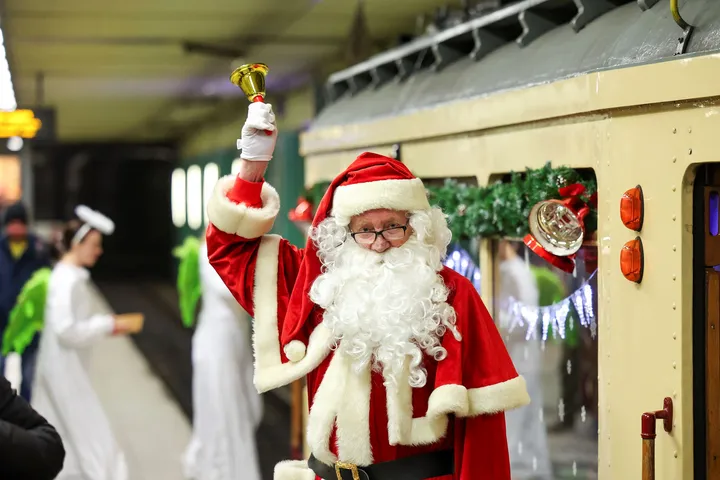 Ein Mann im Nikolauskostüm steht auf dem Bahnsteig einer Tunnelstation vor einer weihanchtlich geschmückten historischen Stadtbahn. In der Hand läutet er eine kleine Glocke.t seinem 