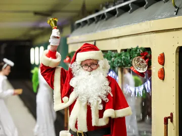 Ein Mann im Nikolauskostüm steht auf dem Bahnsteig einer Tunnelstation vor einer weihanchtlich geschmückten historischen Stadtbahn. In der Hand läutet er eine kleine Glocke.t seinem 