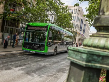 Ein Bus der Linie 100 steht an der Haltestelle Kröpcke vor dem GOP in Hannovers Innenstadt. Der Fotograf steht auf der gegenüberliegenden Straßenseite und blickt hinter einer Steinsäule hervor.
