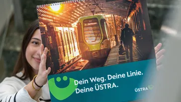 Eine junge Frau hält den ÜSTRA Bildkalender 2026 in die Höhe. Ihr Gesicht ist dabei zur Hälfte bedeckt.