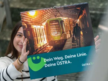 Eine junge Frau hält den ÜSTRA Bildkalender 2026 in die Höhe. Ihr Gesicht ist dabei zur Hälfte bedeckt.