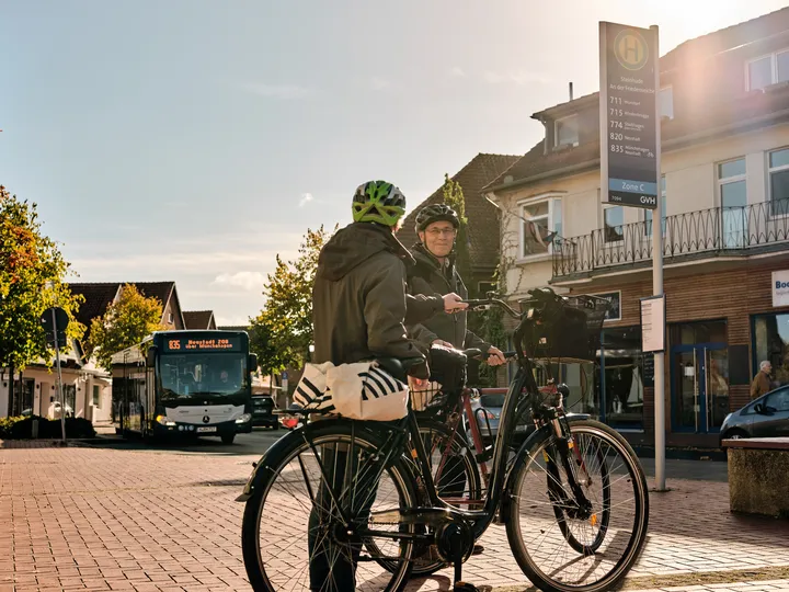 Zwei Menschen stehen mit ihren Fahrrädern an einer Bushaltestelle in Steinhude. Sie tragen Helme und warten auf den Bus, der gerade angefahren kommt. Die Sonne scheint, der Himmel ist blau. 